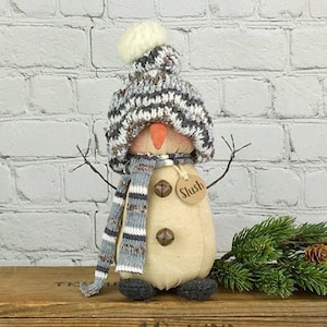 May include: A fabric snowman with a knitted hat and scarf, and a tag that says "Slush". The snowman has a carrot nose, twig arms, and button eyes. It is decorated with a pine branch and a small pinecone.