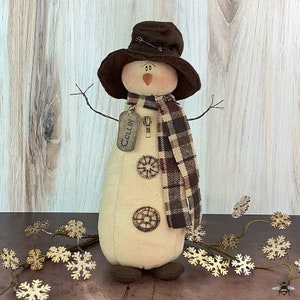 May include: A handcrafted snowman figure with a brown hat, scarf, and boots. The snowman has a tag that says "Ollin". The scarf is a brown and tan plaid pattern. The snowman has wire arms and decorative gear accents.