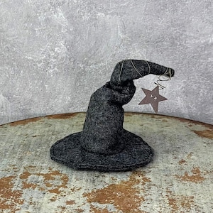 May include: A dark gray felt witch hat with a wide brim and a pointed top. A small, brown star charm hangs from the tip of the hat, suspended by a wire coil. The hat is a whimsical decoration.