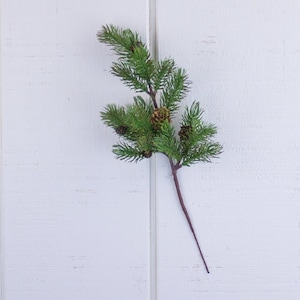 May include: A single sprig of artificial pine with brown pine cones. The pine sprig is green and has a brown stem.