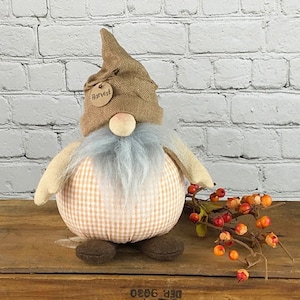 May include: A brown and tan gnome with a gingham patterned body and a burlap hat with a "Harvest" tag. The gnome is sitting on a wooden surface with a sprig of red berries.