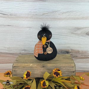 May include: Black crow-shaped handmade doll with a black and orange plaid scarf, a yellow beak, and a brown tag that says "COOP". The doll is sitting on a wooden surface with sunflowers in the foreground.