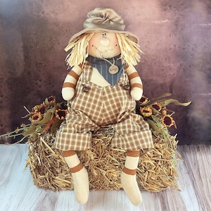 May include: A brown and tan plaid scarecrow doll with a straw hat, a blue bandana, and a name tag that says "Sergio". The scarecrow is sitting on a bale of hay with sunflowers in the background.