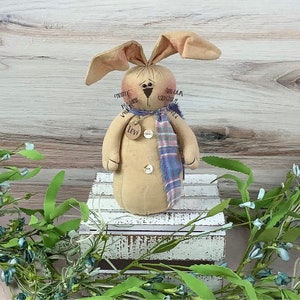 May include: A handmade beige bunny doll with a blue and red plaid scarf. The doll has a button nose and black eyes. The doll is sitting on a white wooden crate. The doll has a tag that says "Levi" on it.