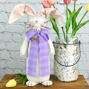 May include: A white bunny doll with long ears and a purple plaid scarf. The doll is wearing a tag that says "fuzzy". The doll is standing in front of a glass jar filled with white paper shreds and a bouquet of yellow and pink tulips.