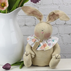May include: A beige fabric bunny with long ears, a pink nose, and black eyes. The bunny is wearing a white ruffled collar with a floral print and a brown tag that says "Dallas".