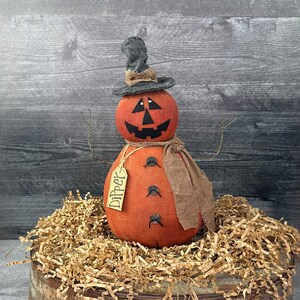 May include: A handcrafted Halloween decoration featuring an orange pumpkin-shaped figure with a carved face, wearing a gray hat and a brown scarf. The figure has wire arms and a tag that says "Dipper". It is displayed on a bed of shredded paper.