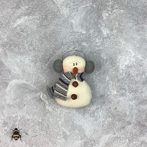 Könnte beinhalten: Ein weißer Schneemann-Anhänger mit einem grauen Schal, grauen Ohrenschützern und braunen Knöpfen. Der Schneemann hat eine rote Nase und schwarze Augen. Der Anhänger befindet sich auf einem grauen Hintergrund.