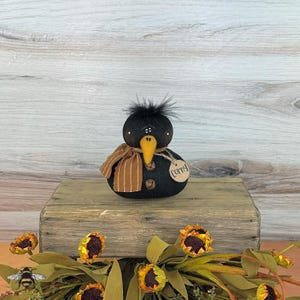 May include: A black crow-like decorative figure with a yellow beak and a brown and tan striped scarf. The figure has a tag that says "Corny". The figure is sitting on a wooden block with sunflowers and greenery in the foreground.