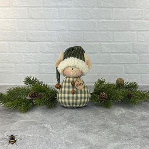 May include: A whimsical elf doll with a green and white plaid body and a matching hat with a white fur trim. The doll has a tag that says "Tinker" and is decorated with small bells. Pine branches and pine cones are arranged around the doll.