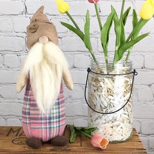 May include: A plaid gnome with a long white beard and a brown hat with the word "Merry" on it. The gnome is standing in front of a glass jar filled with shredded paper and tulips.