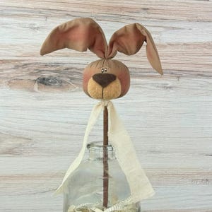 May include: A decorative Easter bunny head on a wooden stick. The bunny has brown and pink fabric ears, a tan nose, and a light brown face. A cream-colored ribbon is tied around the stick. The bunny is placed in a clear glass bottle.