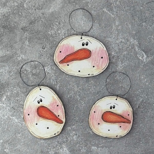 May include: Three snowman ornaments with a hand-drawn style. Each ornament features a white face with pink cheeks, a carrot nose, black dot eyes, and a black wire loop for hanging. The ornaments are on a gray background.
