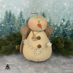May include: A fabric snowman angel with a burlap body, wings, and a halo. The snowman has a peach-colored face with a carrot nose and button eyes. It has a wooden tag that says "Believe" and is decorated with snowflake accents.