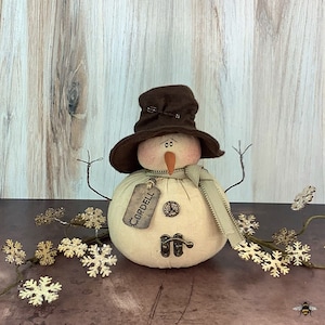 May include: A handmade fabric snowman with a brown felt hat, a zipper mouth, and a metal tag that says "Cordell". The snowman is sitting on a brown surface with gold snowflake decorations.