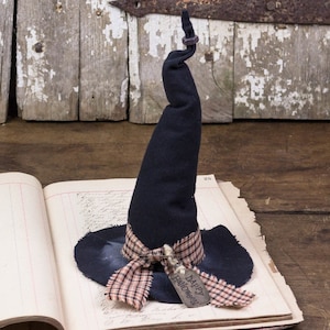 May include: A black witch hat with a pointed top and a brown and orange plaid ribbon tied around the base. The hat is sitting on an open book with the text "Happy Halloween" on a tag attached to the ribbon.