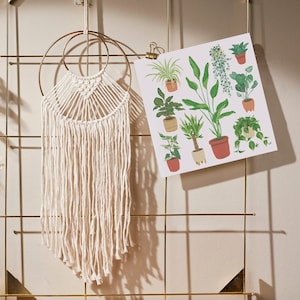 Könnte beinhalten: Ein weißer Makramee-Wandbehang mit einem goldenen Reifen und einem Druck von Topfpflanzen an einer goldenen Gitterwand.