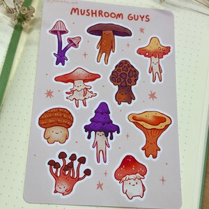 Pode incluir: Uma folha de adesivos com vários personagens de cogumelos de desenho animado em tons de laranja, roxo e marrom. O texto "MUSHROOM GUYS" é exibido no topo. A folha de adesivos está em um fundo rosa claro com estrelas.