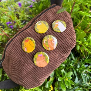 Könnte beinhalten: Fünf bunte Pins mit Tierillustrationen auf einer braunen Cordtasche. Jeder Pin zeigt ein anderes Tier, das eine Blumenkrone trägt: ein Frosch, eine Gans, eine Katze, ein Hund und ein Bär.