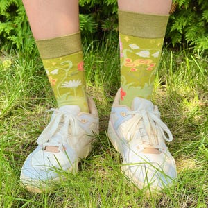 Op de afbeelding: Groene sokken met een bloemenpatroon worden gedragen met witte sneakers, tegen een grasachtige achtergrond. De sokken hebben een lichtgroene basis met een donkerdere groene boord en hebben een ontwerp van bloemen en wolken.