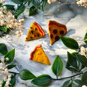 Könnte beinhalten: Drei Miniatur-Pizzascheiben aus Polymer Clay. Die Scheiben sind in verschiedenen Varianten: eine mit Pepperoni, eine mit grünen Paprikaschoten und Oliven und eine nur mit Käse. Die Scheiben sind auf einer weißen Marmorfläche mit grünen Blättern angeordnet.
