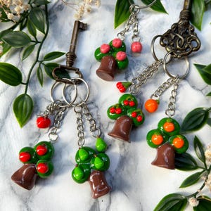 Könnte beinhalten: Schlüsselanhänger mit Miniatur-Baum-Anhängern. Die Anhänger haben braune Baumstämme, grünes Laub und rote oder orangefarbene Früchte. Die Schlüsselanhänger haben silberne Ketten und sind an Schlüsseln im antiken Stil befestigt.