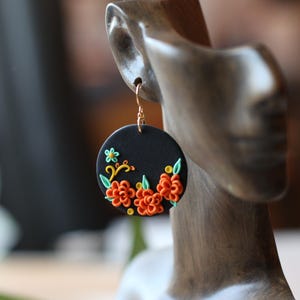 Puede incluir: Un pendiente circular negro con un gancho dorado. El pendiente presenta un dise&ntilde;o floral con detalles en naranja, verde y amarillo. El pendiente se muestra en una cabeza de maniqu&iacute;.