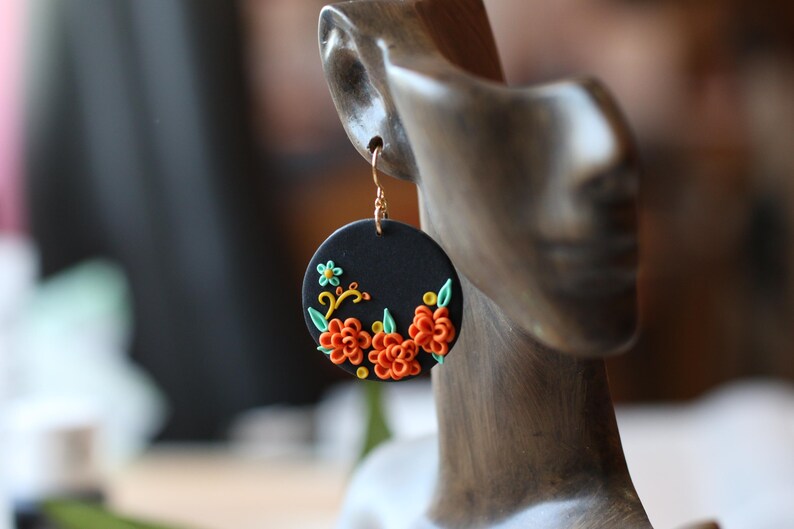 Puede incluir: Un pendiente circular negro con un gancho dorado. El pendiente presenta un dise&ntilde;o floral con detalles en naranja, verde y amarillo. El pendiente se muestra en una cabeza de maniqu&iacute;.