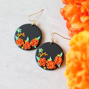 Puede incluir: Un par de pendientes redondos negros con ganchos dorados. Cada pendiente presenta un dise&ntilde;o floral con flores naranjas, hojas verdes y detalles amarillos. Los pendientes est&aacute;n colocados sobre un fondo blanco con flores naranjas en el fondo.
