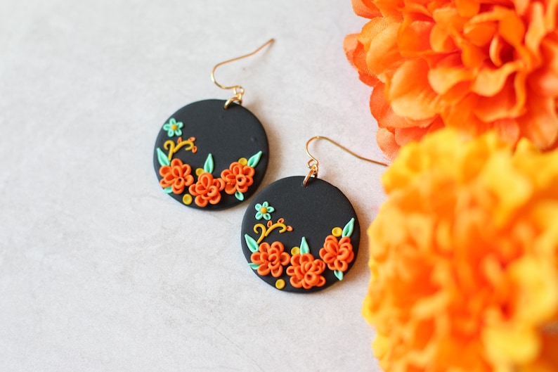 Puede incluir: Un par de pendientes redondos negros con ganchos dorados. Cada pendiente presenta un dise&ntilde;o floral con flores naranjas, hojas verdes y detalles amarillos. Los pendientes est&aacute;n colocados sobre un fondo blanco con flores naranjas en el fondo.