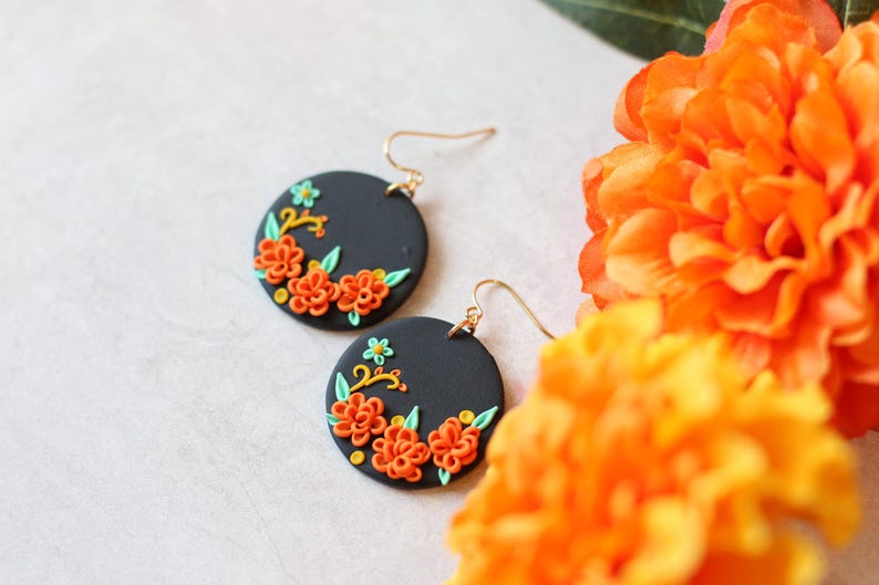 Puede incluir: Un par de pendientes redondos negros con ganchos dorados. Cada pendiente presenta un dise&ntilde;o floral con flores naranjas y amarillas, hojas verdes y peque&ntilde;os detalles turquesas. Los pendientes se muestran cerca de flores naranjas.