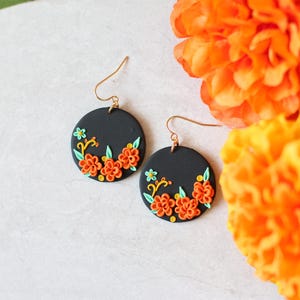 Puede incluir: Un par de pendientes redondos negros con ganchos dorados. Cada pendiente presenta un dise&ntilde;o floral con flores naranjas y amarillas, hojas verdes y detalles amarillos. Los pendientes se muestran con flores de color naranja brillante en el fondo.