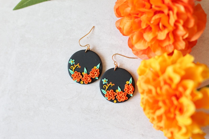 Puede incluir: Un par de pendientes redondos negros con ganchos dorados. Cada pendiente presenta un dise&ntilde;o floral con flores naranjas y amarillas, hojas verdes y detalles amarillos. Los pendientes se muestran con flores de color naranja brillante en el fondo.