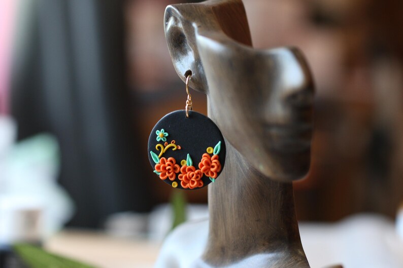 Puede incluir: Pendientes circulares negros con dise&ntilde;os florales naranjas y amarillos. Los pendientes tienen un gancho dorado y se exhiben en una cabeza de maniqu&iacute; de madera. Las flores tienen hojas verdes.