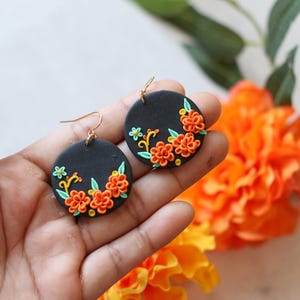 Puede incluir: Pendientes redondos negros con dise&ntilde;os florales naranjas y amarillos. Los pendientes tienen ganchos dorados. Los pendientes se sostienen en una mano, con flores naranjas en el fondo.