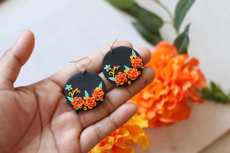 Puede incluir: Pendientes redondos negros con dise&ntilde;os florales naranjas y amarillos. Los pendientes tienen ganchos dorados. Los pendientes se sostienen en una mano, con flores naranjas en el fondo.