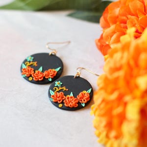 Puede incluir: Un par de pendientes redondos negros con ganchos dorados. Cada pendiente presenta un dise&ntilde;o floral con flores naranjas, hojas verdes y detalles amarillos. Los pendientes se exhiben cerca de flores de color naranja brillante.