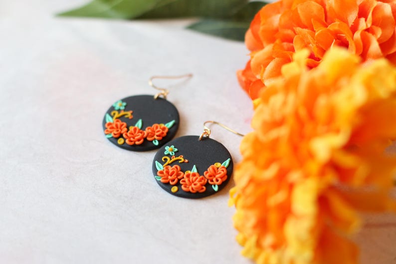 Puede incluir: Un par de pendientes redondos negros con ganchos dorados. Cada pendiente presenta un dise&ntilde;o floral con flores naranjas, hojas verdes y detalles amarillos. Los pendientes se exhiben cerca de flores de color naranja brillante.