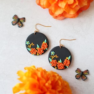 Puede incluir: Pendientes redondos negros con dise&ntilde;os florales naranjas y amarillos y ganchos dorados. Tambi&eacute;n hay dos peque&ntilde;os adornos en forma de mariposa. Los pendientes est&aacute;n colocados sobre un fondo blanco con flores naranjas.