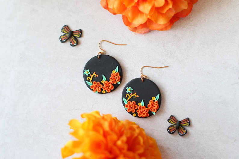 Puede incluir: Pendientes redondos negros con dise&ntilde;os florales naranjas y amarillos y ganchos dorados. Tambi&eacute;n hay dos peque&ntilde;os adornos en forma de mariposa. Los pendientes est&aacute;n colocados sobre un fondo blanco con flores naranjas.