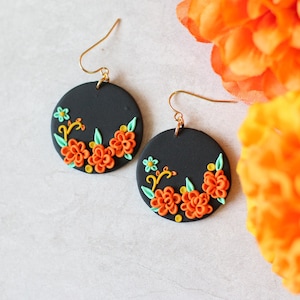 Puede incluir: Un par de pendientes redondos negros con ganchos dorados. Cada pendiente presenta un dise&ntilde;o floral con flores naranjas, hojas verdes y detalles amarillos. Los pendientes est&aacute;n sobre un fondo blanco con flores naranjas.