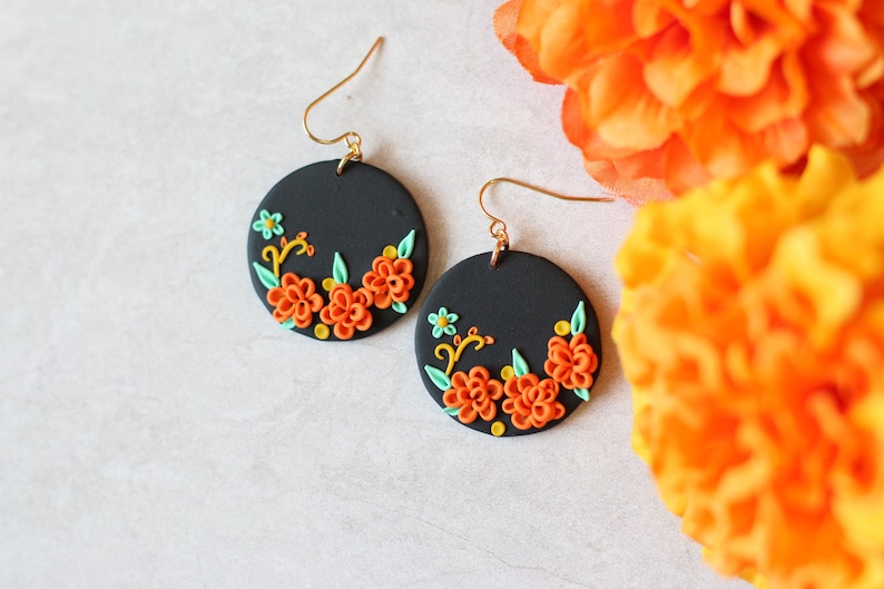 Puede incluir: Un par de pendientes redondos negros con ganchos dorados. Cada pendiente presenta un dise&ntilde;o floral con flores naranjas, hojas verdes y detalles amarillos. Los pendientes est&aacute;n sobre un fondo blanco con flores naranjas.