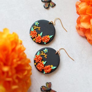 Puede incluir: Un par de pendientes redondos negros con dise&ntilde;os florales naranjas y verdes. Los pendientes tienen ganchos dorados. La imagen tambi&eacute;n presenta flores naranjas y una decoraci&oacute;n en forma de mariposa.