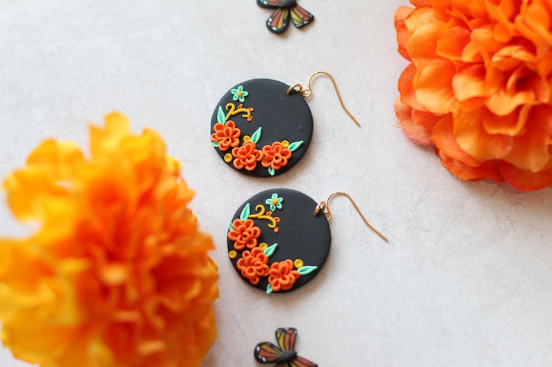 Puede incluir: Un par de pendientes redondos negros con dise&ntilde;os florales naranjas y verdes. Los pendientes tienen ganchos dorados. La imagen tambi&eacute;n presenta flores naranjas y una decoraci&oacute;n en forma de mariposa.