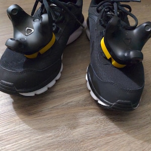 May include: A pair of black sneakers with white soles and yellow accents. Each shoe has a black VR controller attached to the side with a yellow strap.