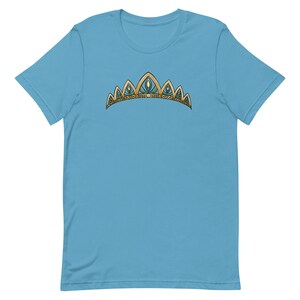 May include: A light blue t-shirt with a gold and green crown design. The crown has five points and is decorated with small, round, blue gems.