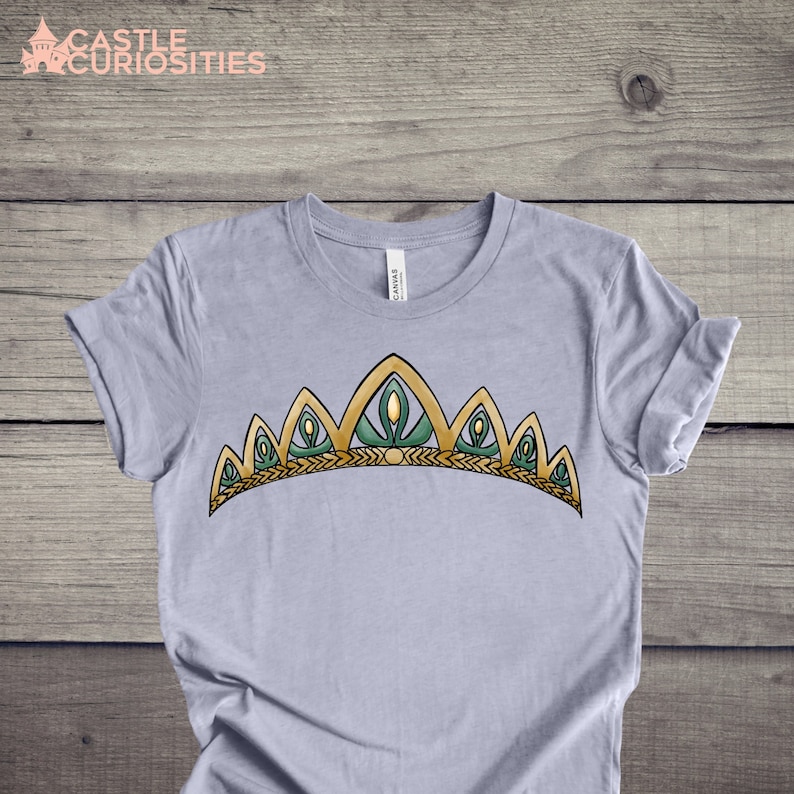 May include: A light gray t-shirt with a gold and green crown design. The crown has a pointed top and a row of green leaves along the bottom.