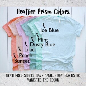 May include: A set of six heather t-shirts in various pastel colors, including peach, sunset, lilac, dusty blue, mint, and ice blue. The shirts are arranged in a row from darkest to lightest, with the text "Heather Prism Colors" at the top and "HEATHERED SHIRTS HAVE SMALL GREY FLECKS TO VARIGATE THE COLOR" at the bottom.