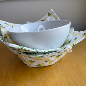 Puede incluir: Un cuenco de cerámica blanco anidado en un salvabols de patrón de limón. El salvabols es blanco con limones amarillos y hojas verdes. El cuenco y el salvabols están sobre una superficie de madera marrón claro.