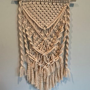 May include: A beige macrame wall hanging with a geometric pattern and fringe. The hanging is made with natural cotton cord and is attached to a wooden dowel.